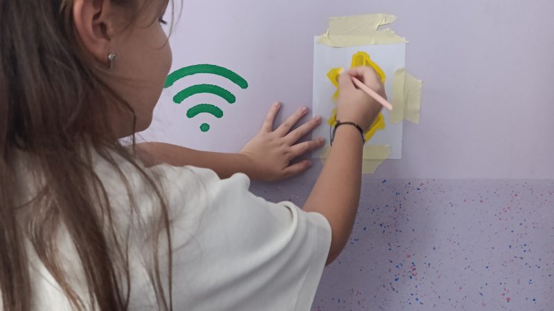Student painting a yellow icon on the classroom wall with a stencil next to a green WiFi symbol